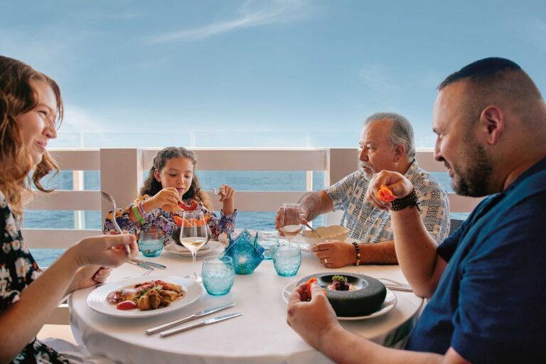 A family enjoys a fine dining experience on the Carnival Mardi Gras cruise, with an ocean view in the background, as they savor gourmet dishes and share a joyful meal together.