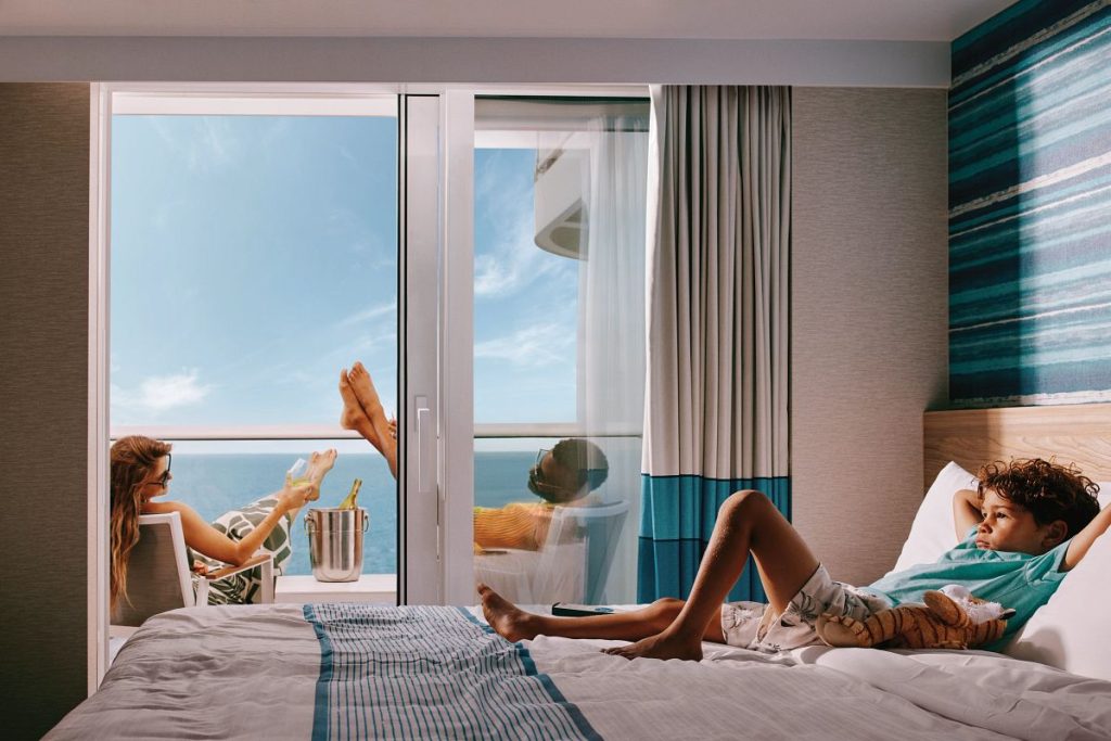 A relaxing moment captured in a Carnival cruise balcony room where a woman enjoys a glass of champagne on the balcony while a child lounges on the bed, with the tranquil sea visible through the open glass door.