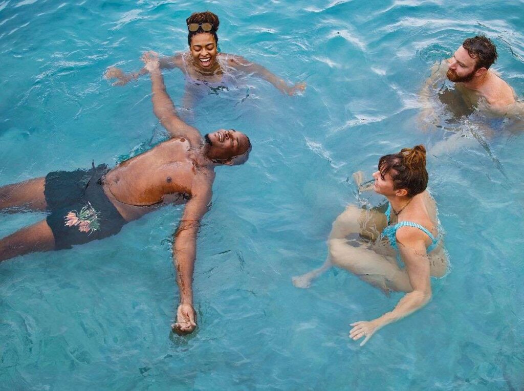 The image depicts a group of cruise guests engaged in a water workout in the ship's pool, each with cheerful expressions and enjoying the activity. The setting suggests a fun, communal atmosphere where passengers can participate in fitness activities while socializing and enjoying the onboard amenities.