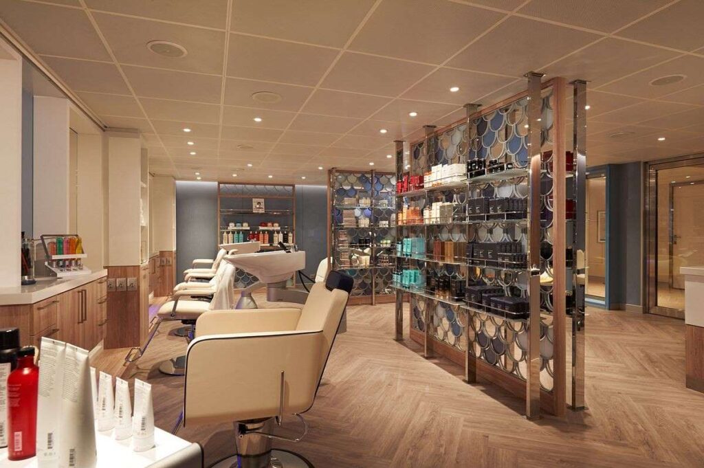 The image showcases the modern and stylish interior of the Beauty Salon at Carnival Sunrise's Cloud 9 Spa. It features a row of salon chairs facing a mirror-lined wall, hair care stations equipped with professional styling tools, and a vast array of beauty products neatly displayed on glass shelving. The elegant design and warm lighting create a relaxing atmosphere for guests seeking pampering services.