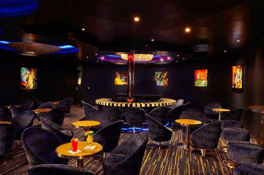 Piano Bar 88 on Carnival Sunrise exudes a cool, jazzy ambiance with plush velvet chairs, vibrant artwork on the walls, and a sleek grand piano set against a backdrop of soft blue lighting.