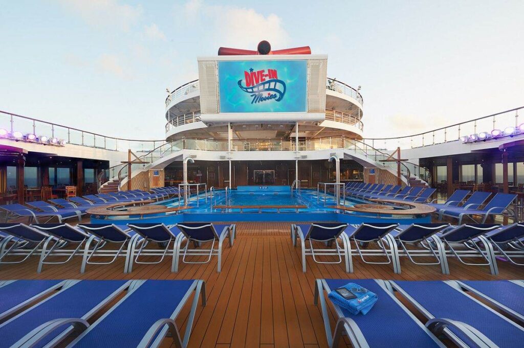 Carnival Sunrise's pool deck set up for Dive-In Movies, featuring rows of blue loungers facing a large outdoor screen, with a tranquil pool reflecting the early evening sky.