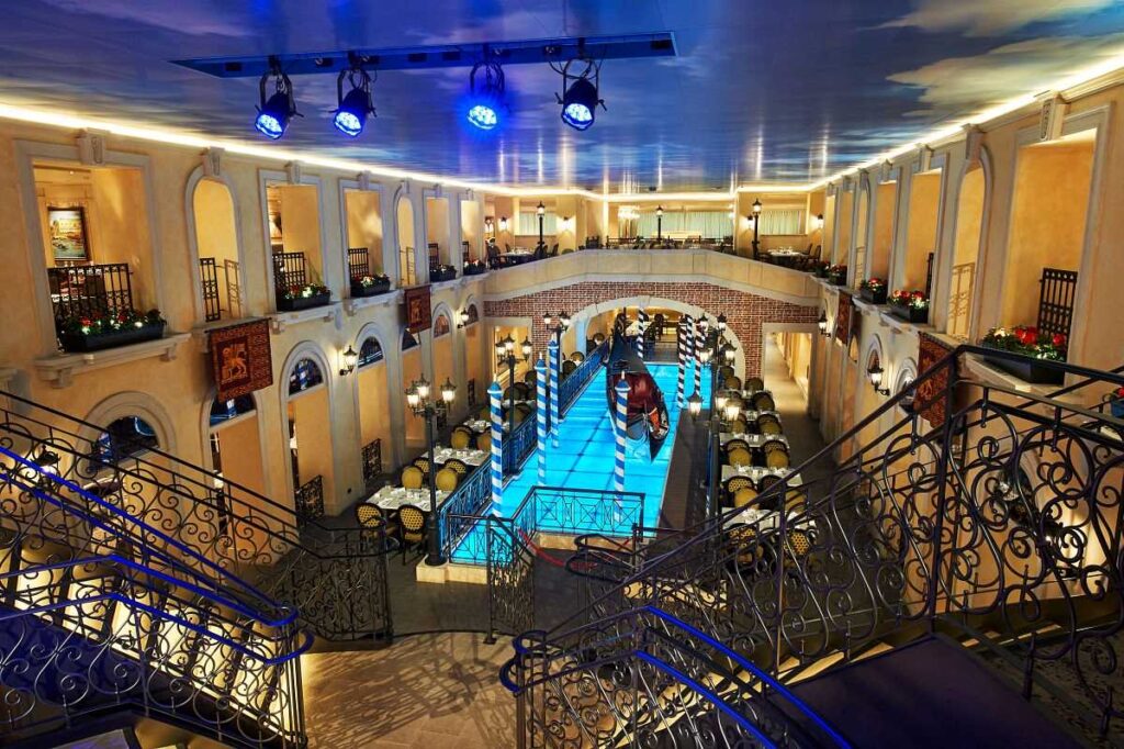 Elevated view of the Canal Grande restaurant inside the Carnival Venezia cruise ship, showcasing its Venetian-inspired architecture, with a central blue-lit canal, gondola, and surrounding dining areas adorned with festive decorations.