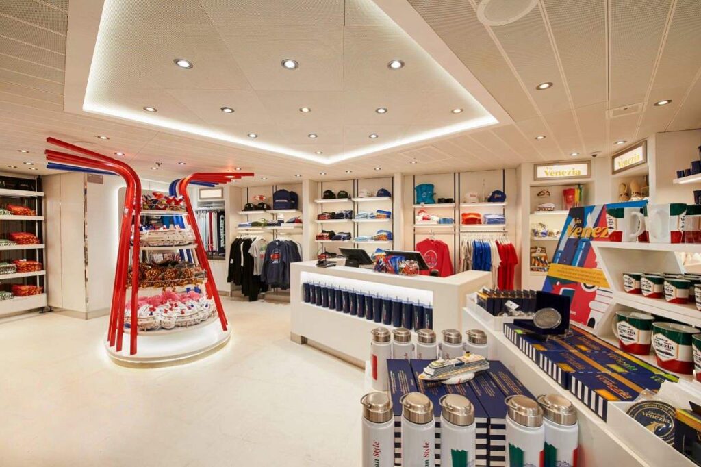 The image captures the interior of the Carnival Venezia store, showcasing a bright and neatly organized retail space with a variety of souvenirs and apparel on display. The color theme of red, white, and blue, along with the sleek shelving and spacious design, provides a welcoming shopping environment for cruise guests.