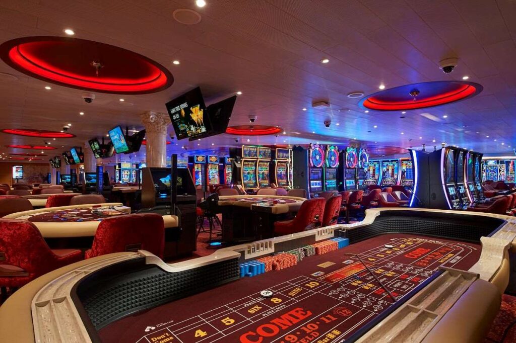The casino on Carnival Venezia, stylishly designed with bright red accents from circular light fixtures overhead. The space is filled with a variety of gaming options including tables for card games and a selection of slot machines, all set against a backdrop of plush red chairs and vibrant carpeting, creating an exciting and dynamic environment for guests to enjoy.