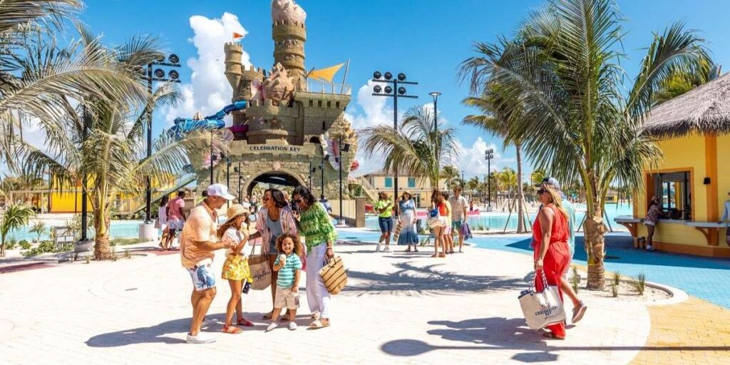 Families stroll under palm trees at Celebration Key, a tropical-themed cruise destination featuring a large sandcastle play structure with colorful water slides and a sign reading "Celebration Key." Guests are dressed in beachwear, enjoying the sun-drenched setting surrounded by pools, yellow cabanas, and island-style huts.