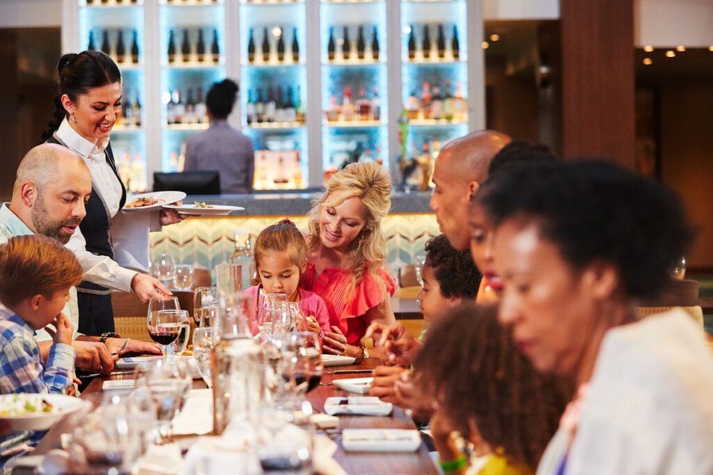 A lively dining scene in the main dining room on Carnival Vista, where a waiter serves dishes to a family at a table, with children looking excited, a wine bottle being poured, and a backdrop of a well-stocked bar.