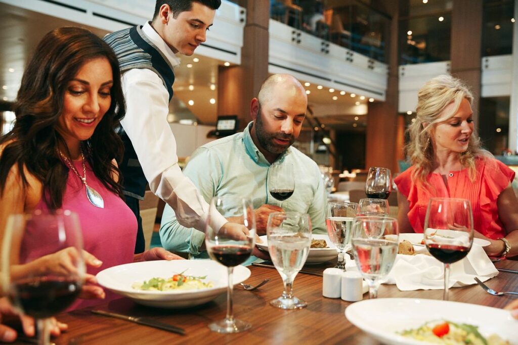This image shows a group of Carnival cruise guests enjoying a fine dining experience. A server in a vest is attending to the table, delivering dishes to a couple seated among elegantly set plates, wine glasses, and water. The warm lighting and stylish restaurant ambiance emphasize the luxurious and social dining atmosphere on the cruise.