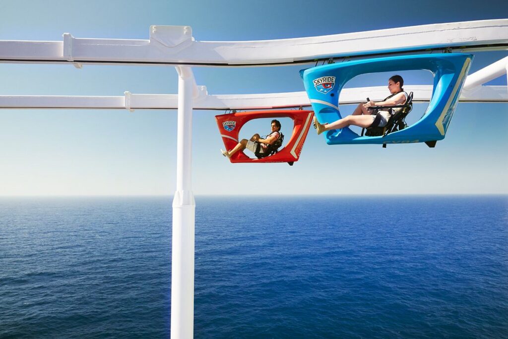 Passengers enjoy the SkyRide attraction aboard the Carnival Vista, pedaling in suspended colorful bike cars against the endless blue of the ocean and sky.