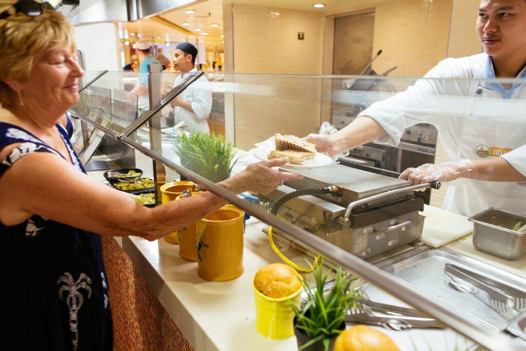 A woman receives a freshly prepared sandwich from a chef at the Deli in the Marketplace on the Carnival Breeze cruise ship. The deli counter features a variety of fresh ingredients and is decorated with small potted plants, adding a touch of greenery. The chef, wearing a white uniform and gloves, hands the plate over the glass partition, ensuring a hygienic and appetizing service experience.