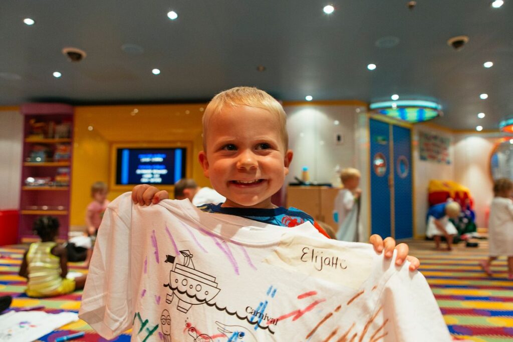 A joyful young boy at Camp Carnival's Night Owls, proudly holding up a white T-shirt with colorful hand-drawn designs, with other children engaged in activities in the background.