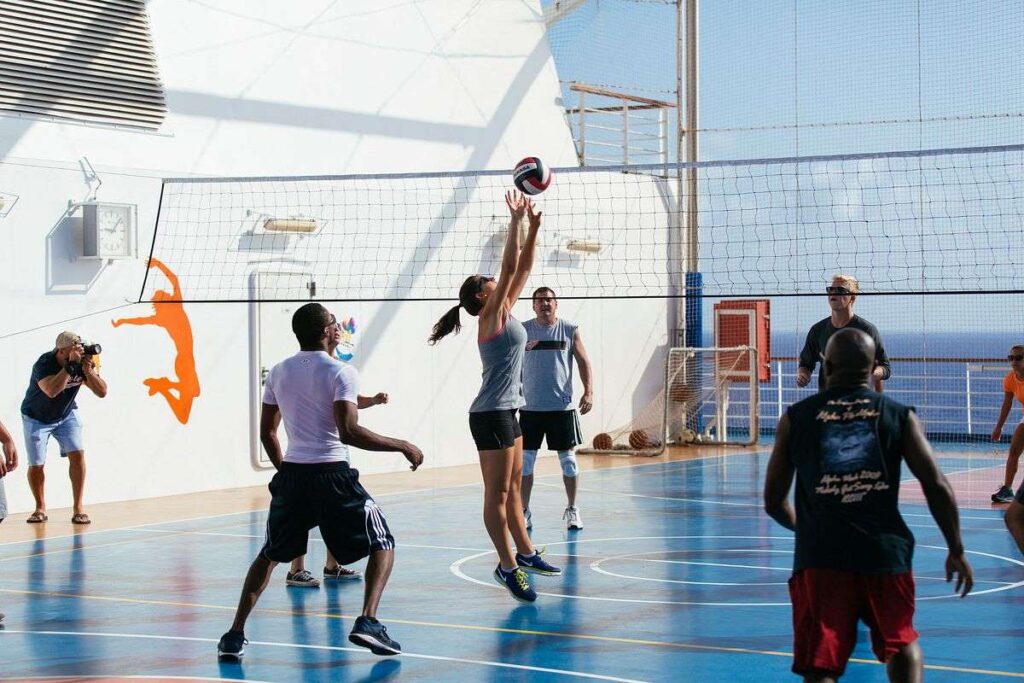 The image showcases an active volleyball game at the SportSquare on the Carnival Breeze, with passengers fully engaged in the match. The outdoor court is situated high on the deck with protective netting around, ensuring a safe environment for a spirited game against the backdrop of the open sea and sky. The scene is a testament to the recreational activities available on the cruise, catering to fitness enthusiasts and sports fans alike.
