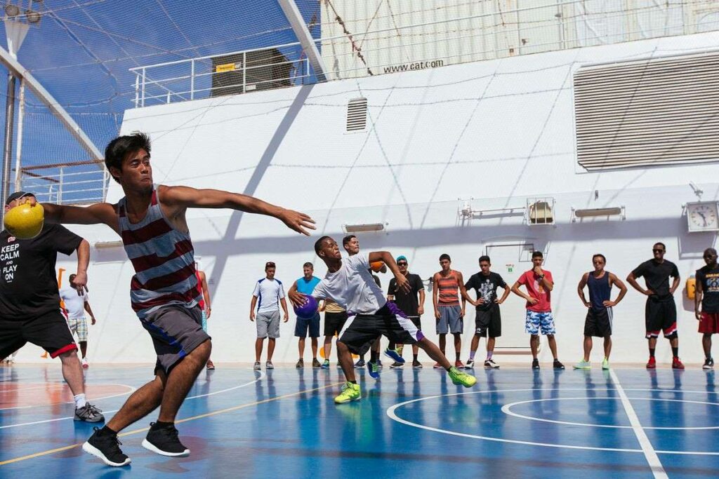 The image depicts an intense game of dodgeball at the SportSquare on the Carnival Breeze, with participants actively engaged in the play. The outdoor court is surrounded by netting against the backdrop of the ship's structure, providing a dynamic setting for recreational activities at sea. The players, in various athletic poses, capture the excitement and competitive spirit of the game.