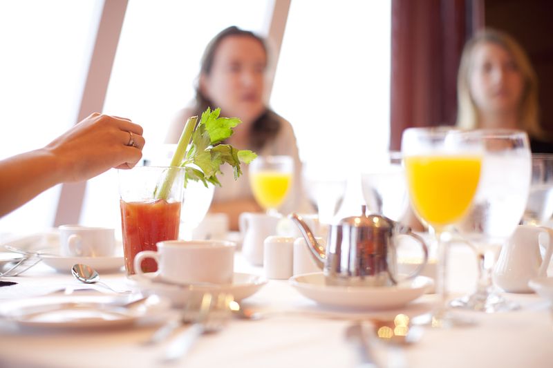 A breakfast table on a Carnival cruise ship, featuring a Bloody Mary garnished with celery, coffee cups, orange juice, and a teapot. Two women are in the background, slightly out of focus, creating a casual and social atmosphere.
