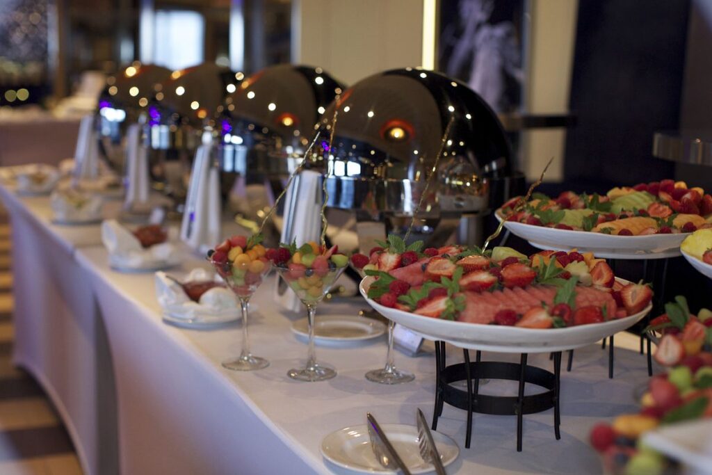 A lavish breakfast buffet is set up on a Carnival cruise ship, featuring a variety of fresh fruits elegantly displayed in large bowls and individual glasses. The buffet table is lined with polished chafing dishes, reflecting the soft lighting of the dining area. The spread exudes an inviting and luxurious atmosphere, perfect for guests to start their day with a delicious and vibrant meal.