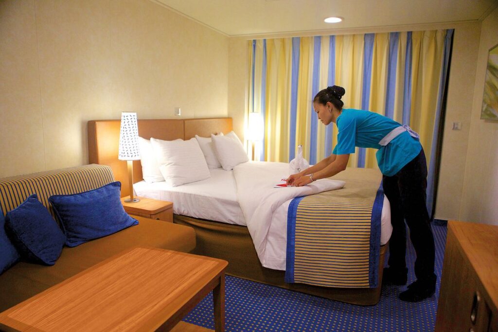 The image shows a room steward in a blue uniform meticulously preparing a towel animal on a neatly made bed in a Carnival Breeze stateroom. The cozy and well-appointed room, with its warm lighting and inviting decor, offers a glimpse into the attentive service and charming touches that enhance the cruise experience for guests.