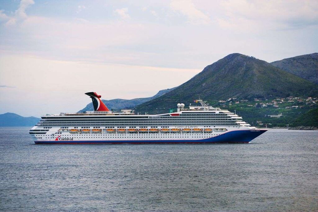 Carnival Liberty cruise ship sails near a mountainous coastline, its signature red, white, and blue funnel marking its presence against the natural landscape. The ocean is calm, reflecting the overcast sky, as the ship adds a touch of human innovation to the serene, natural setting.