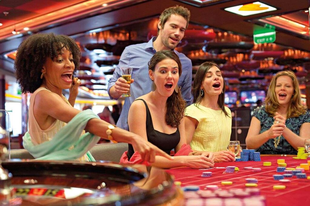 A group of friends at a roulette table in the casino on Carnival Breeze, visibly excited and engaged in the game. They are sharing a fun moment, with expressions of joy and anticipation, drinks in hand, as they participate in the lively and vibrant casino atmosphere.