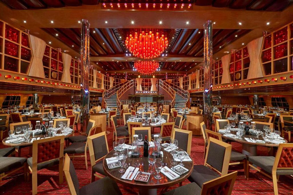 The Crimson Dining Room on the Carnival Dream, set for dinner with a luxurious red and gold color scheme. The room features elegantly set round tables with sparkling glassware and a vibrant red glass chandelier as a centerpiece, reflecting an upscale and welcoming atmosphere for dining.