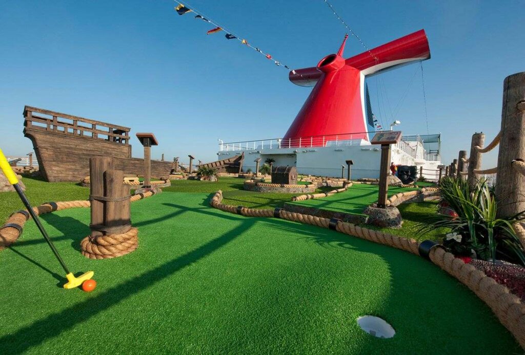 Passengers on the Carnival Dream enjoy a round of mini-golf on a creatively designed course with lush green turf, nautical-themed obstacles, and the ship's iconic red funnel in the background.