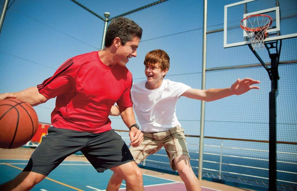 The photograph captures a father and son enjoying a game of basketball on a Carnival cruise ship, sharing a playful and bonding moment. The outdoor court, surrounded by a protective net, provides a perfect setting for active family fun against the backdrop of the open sea, highlighting the recreational amenities offered on the cruise.