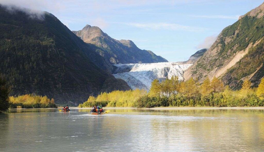 The image captures a scenic Alaskan adventure, a part of Carnival Cruise's shore excursions in Skagway, Alaska. It features a tranquil river with groups of adventurers paddling in inflatable rafts, surrounded by the majestic beauty of towering mountains and a stunning glacier in the distance. The lush greenery of the riverside foliage adds a pop of color to the serene landscape, illustrating a perfect blend of exhilarating activity and breathtaking nature.