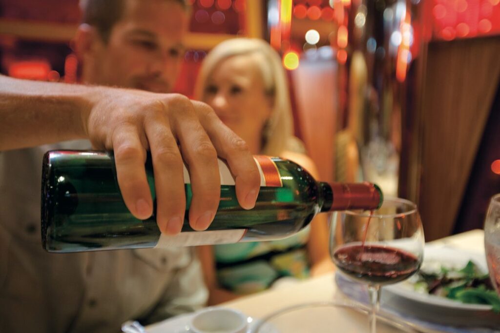 This image depicts a couple enjoying a dining experience aboard a Carnival cruise ship. A hand is seen pouring red wine into a glass, with a blurred background showcasing a warm, inviting dining ambiance. The scene captures the elegance and enjoyment of cruise ship dining, emphasizing relaxation and indulgence.