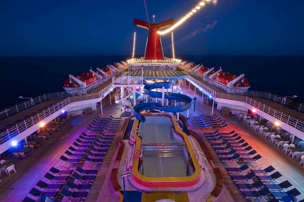 The photo captures the Lido deck of the Carnival Elation at dusk, showcasing the pool area lit by ambient lights with a series of deck chairs lined up for relaxation and the ship's iconic red funnel glowing in the background. It conveys a tranquil evening atmosphere on the cruise.