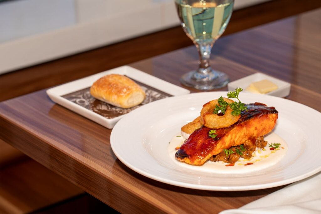 Gourmet BBQ salmon garnished with fresh herbs and a crispy onion ring, accompanied by a golden pastry and a glass of white wine on a polished wooden table in Carnival Cruise's main dining room.