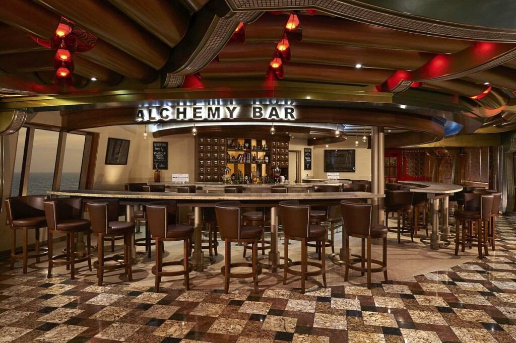 The Alchemy Bar on Carnival Freedom with a sleek and modern design, featuring high stools at the bar counter and a spacious seating area. The interior boasts a mix of dark wood tones, checkerboard floor patterns, and red accent lighting, creating an inviting ambiance for enjoying crafted cocktails at sea.