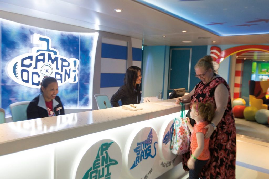 A smiling child and a woman interacting with staff at the colorful 'Camp Ocean' kids club reception on a cruise ship, with vibrant marine-themed decor in the background.
