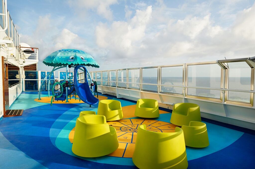 The outdoor play area of Camp Ocean on Carnival Freedom, featuring a playful blue jellyfish-themed water feature with a slide, surrounded by bright yellow seating. The area is designed with a colorful, ocean-inspired floor, and the vast ocean provides a serene backdrop, perfect for children's playtime at sea.