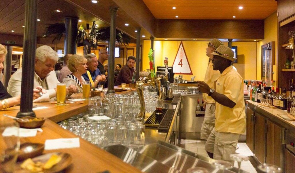 Patrons enjoying the lively atmosphere of the RedFrog Pub on Carnival Freedom, with some seated at the bar enjoying beers and casual conversation. A bartender in a straw hat and yellow shirt is engaging with guests, amidst the warm ambiance highlighted by wood tones and a vibrant pub setting.