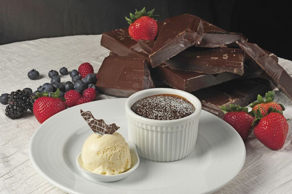Carnival Cruise's signature Chocolate Melting Cake served on a white plate, accompanied by a scoop of vanilla ice cream and garnished with fresh strawberries, blueberries, and blackberries, with chocolate pieces in the backdrop.