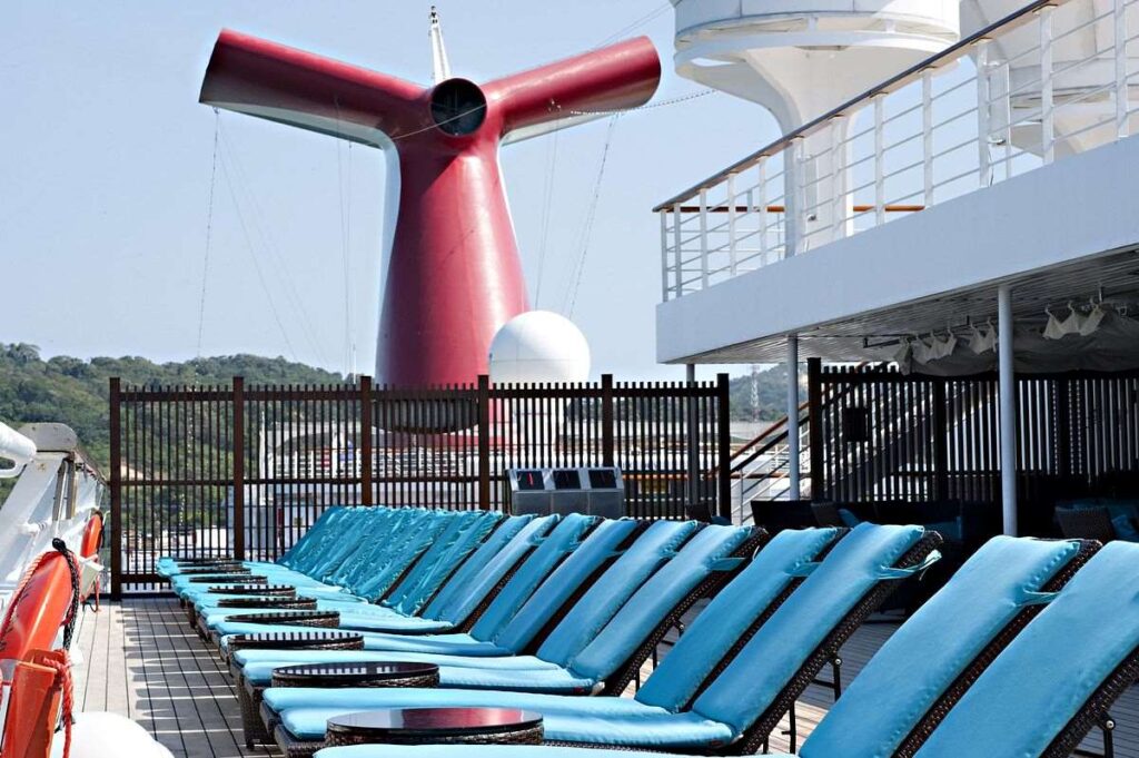 A row of turquoise sun loungers awaits guests on the Serenity deck of the Carnival Glory, with the ship's distinctive red funnel rising in the background.