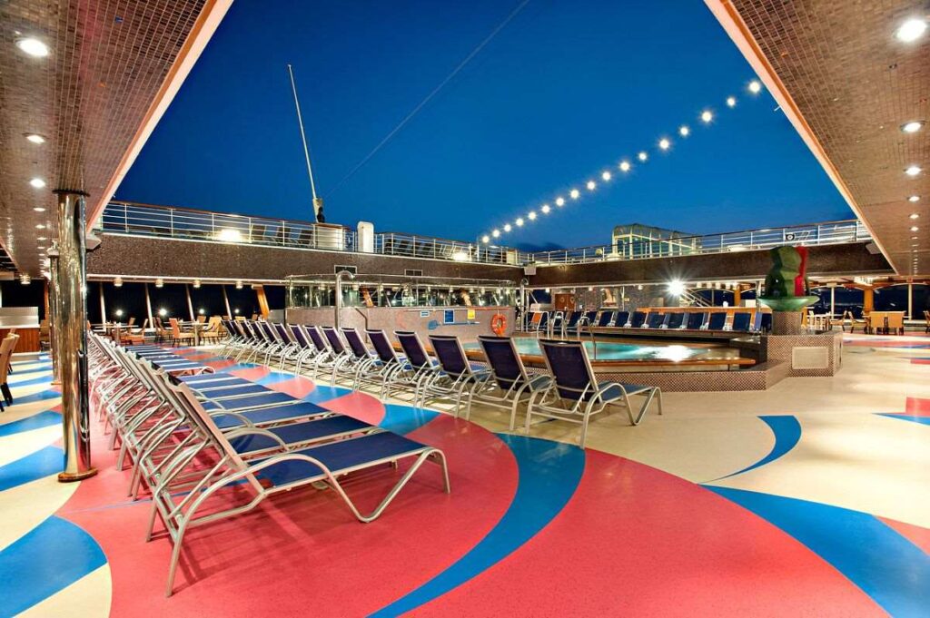 A tranquil evening at the Carnival Glory's Azure Pool, featuring rows of unoccupied loungers ready for guests, with a vibrant patterned deck and string lights adding to the inviting ambiance.