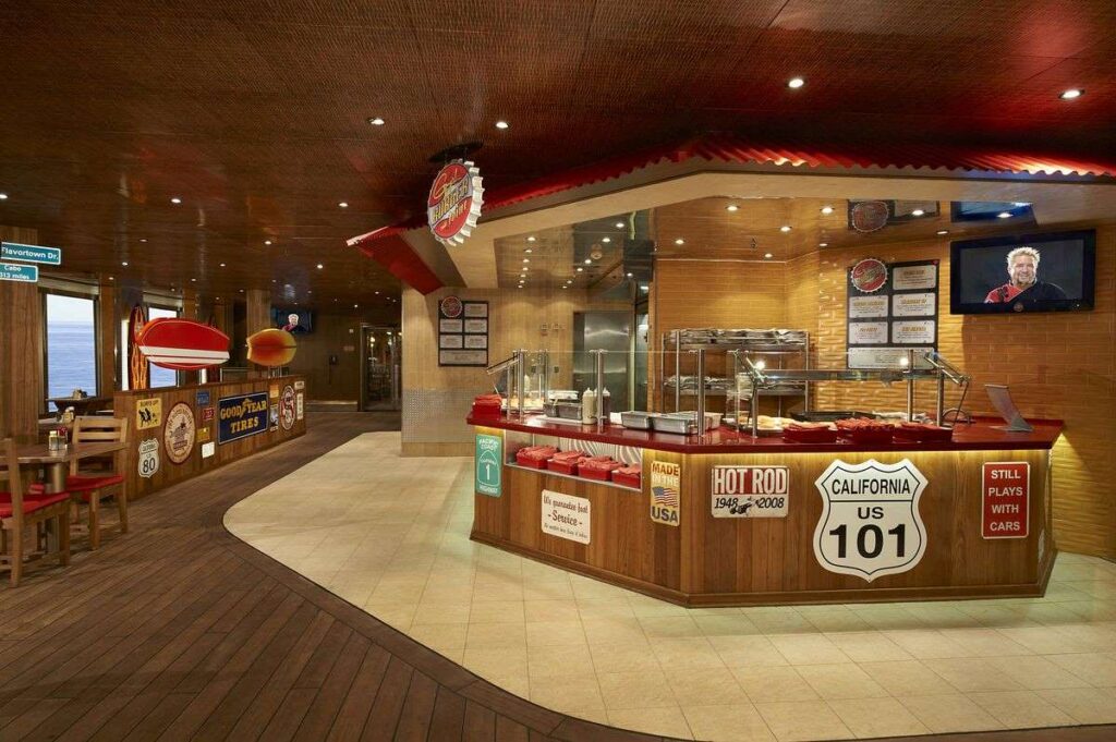 Guy's Burger Joint on Carnival Liberty, featuring a classic American diner theme with a warm wooden interior and nostalgic decor. The serving counter is adorned with signage celebrating the "Hot Rod" and "California US 101," evoking the spirit of vintage car culture, while guests can enjoy watching TV as they wait for their freshly grilled burgers.