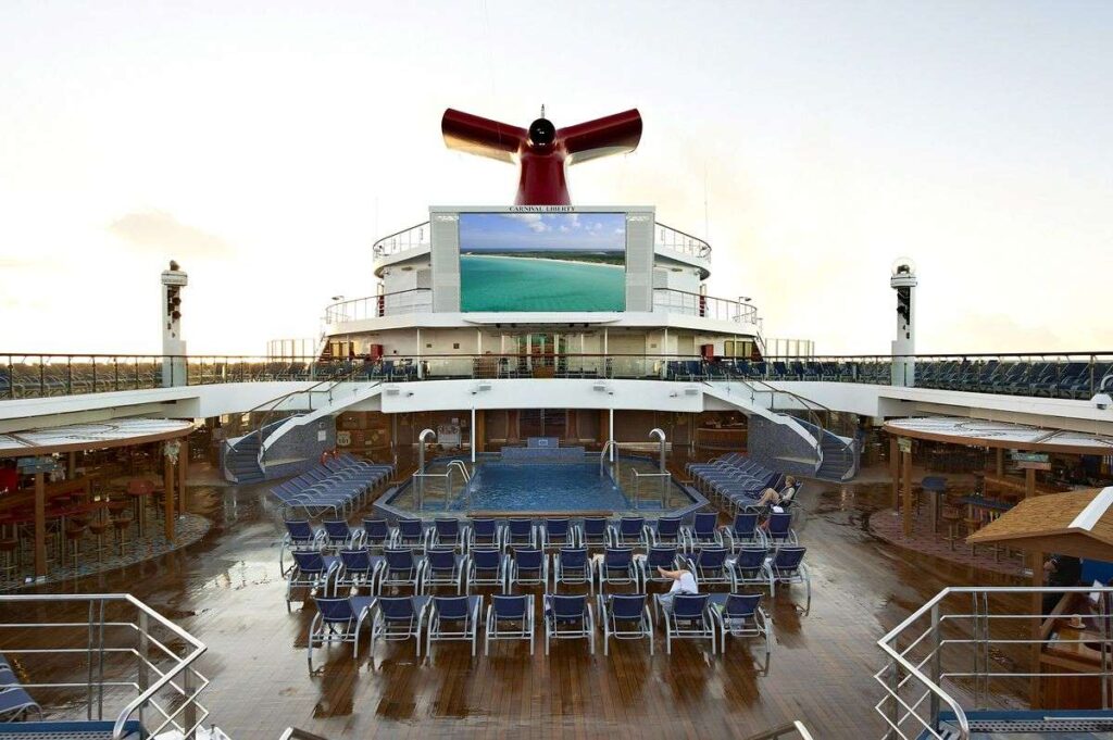 The Seaside Theatre on Carnival Liberty, with its large outdoor screen prominently displaying vivid imagery. The theater is situated on the deck with rows of loungers facing the screen, ready for passengers to relax and enjoy a movie by the pool under the open sky, with the ship's iconic red funnel overhead.