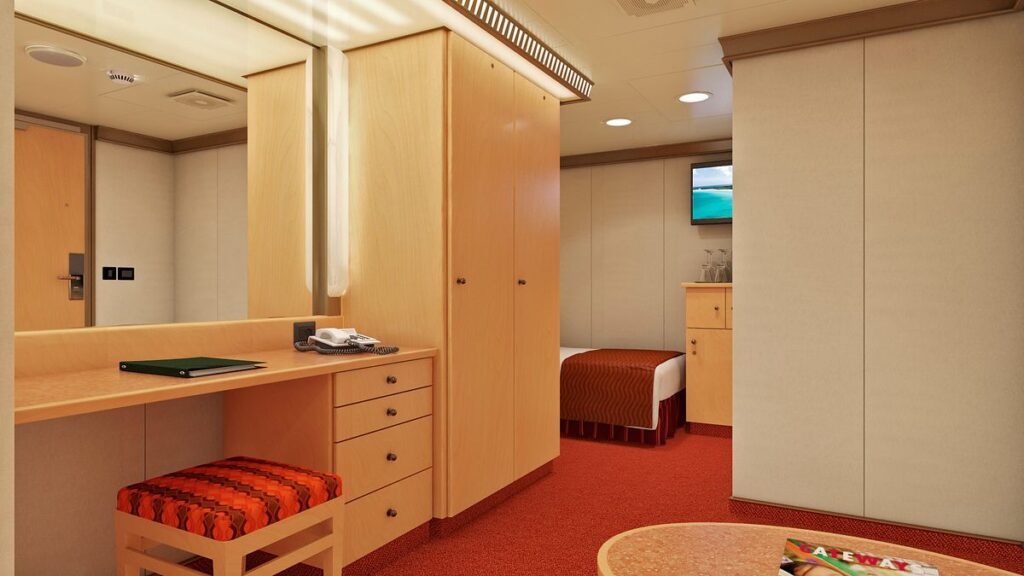 This image shows a stateroom aboard the Carnival Magic cruise ship. The room features light wooden furnishings, including a desk with a phone and a cushioned stool. A bed with a brown blanket is partially visible in the background. The room also includes a small cabinet with glasses on top and a flat-screen TV mounted on the wall. The décor is simple and functional, with soft lighting and a warm, orange-toned carpet.