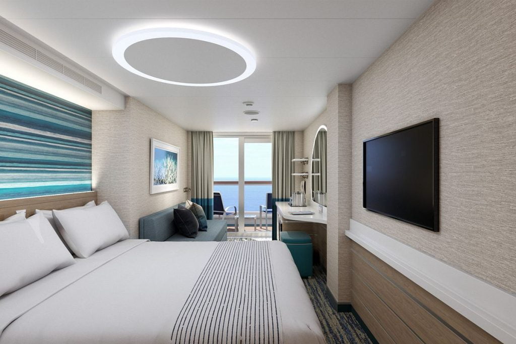 A contemporary Carnival cruise ship junior balcony room featuring a cozy bed, vibrant artwork on the wall, a flat-screen TV, and a sliding door leading to a private balcony with ocean views.