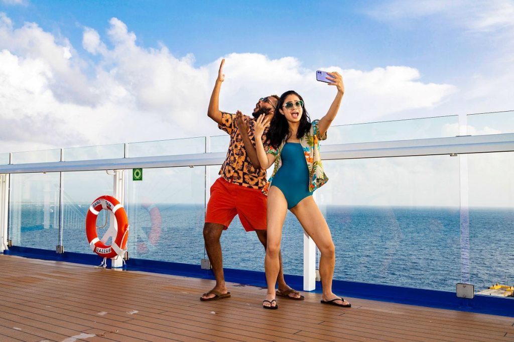A couple takes a selfie on a Carnival cruise ship, with the man playfully raising his hand and the woman posing confidently, both in bright tropical attire against the serene backdrop of the open sea.