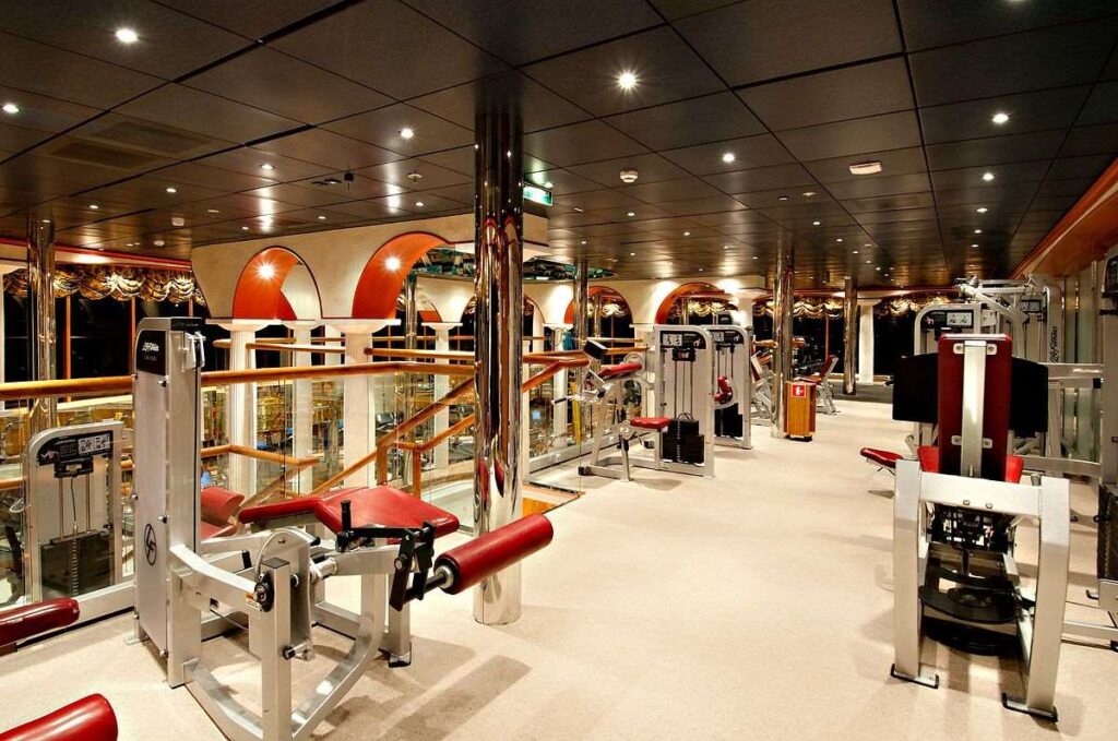 The photograph showcases the well-equipped gym aboard the Carnival Miracle, featuring a variety of weight machines and workout stations. The gym's interior is spacious and mirrored, reflecting the warm lighting and elegant decor, which creates an inviting atmosphere for guests to engage in physical fitness with views of the surroundings through the adjacent windows.
