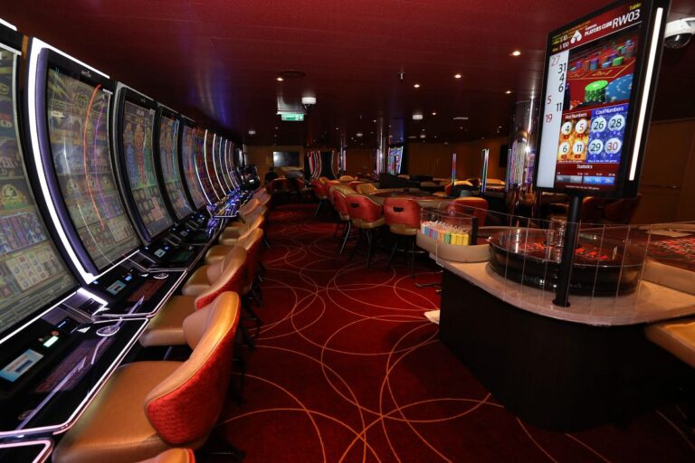 A non-smoking Carnival cruises casino annex featuring a row of modern slot machines on the left with glowing LED accents and individual chairs. In the center and right are several round gaming tables with red and beige chairs, a roulette wheel, and a screen showing betting statistics and "Players Club RW03." The carpet is deep red with circular golden patterns, and the lighting is dim and ambient.