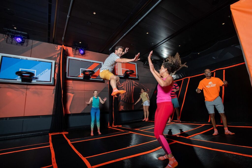 Energetic guests enjoy the Sky Zone Trampoline Park aboard the Carnival Panorama, with a man mid-jump reaching for a high five and others dancing and jumping around him, all surrounded by vibrant orange accents.