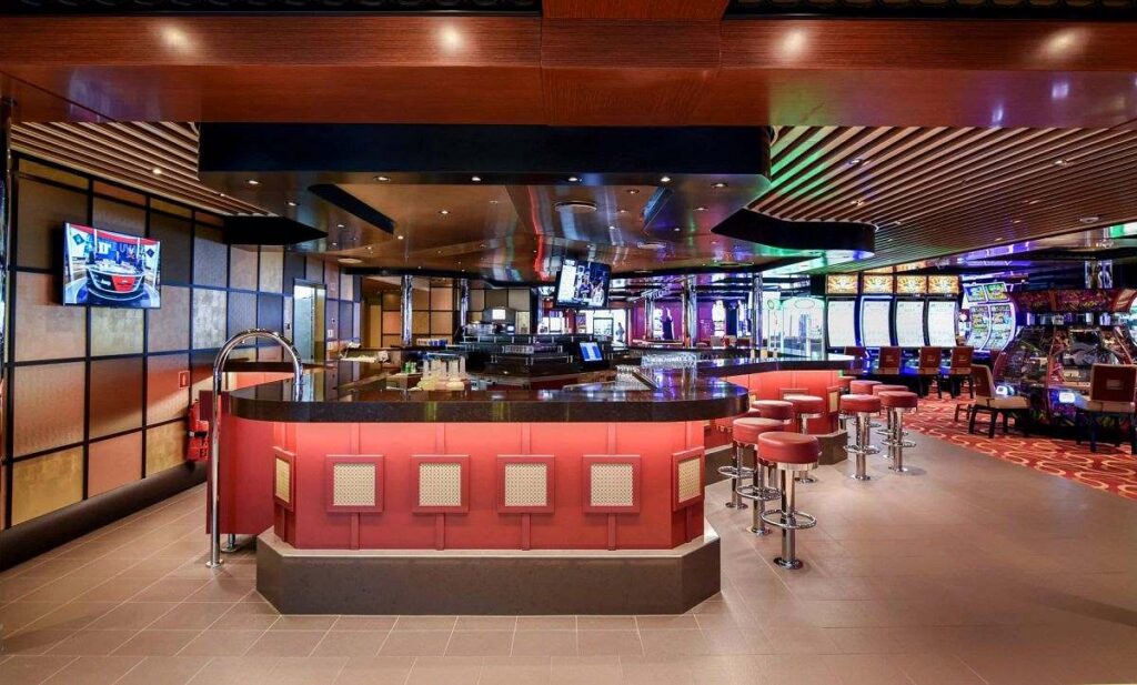 The Casino Bar on Carnival Radiance, displaying a sleek and modern design with a red and black color scheme. The bar features contemporary stools and a polished counter, with gaming machines in close proximity, offering a convenient spot for guests to grab a drink between games in the bustling casino atmosphere.