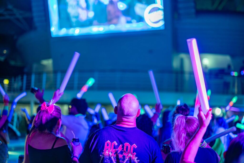 A crowd of people waves glowing foam sticks during the "80s Rock-N-Glow" party on a Carnival Cruise, set against a vibrant, neon-lit deck. The central figure wears an AC/DC "Black Ice" shirt while facing a large screen showing music videos, adding to the retro rock vibe.