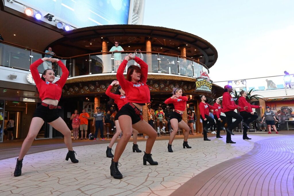 A dance crew in matching red hoodies and black outfits performs a synchronized routine during the Carnival Cruise Tailgate Party, held in front of the RedFrog Tiki Bar. Guests watch from both levels of the deck as performers bring energy to the open-air venue.