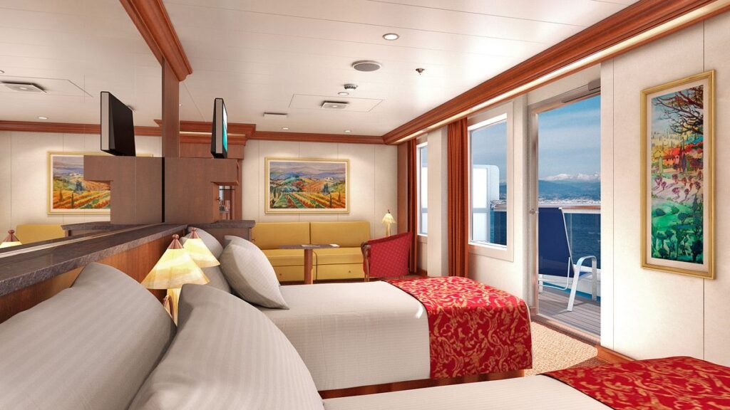 The Ocean Suite on Carnival Freedom, elegantly appointed with two beds with red-patterned bedspreads, a cozy sitting area with a sofa, and a private balcony offering stunning sea views. The room is adorned with framed artwork and warm wooden accents, creating a luxurious and comfortable retreat for passengers.