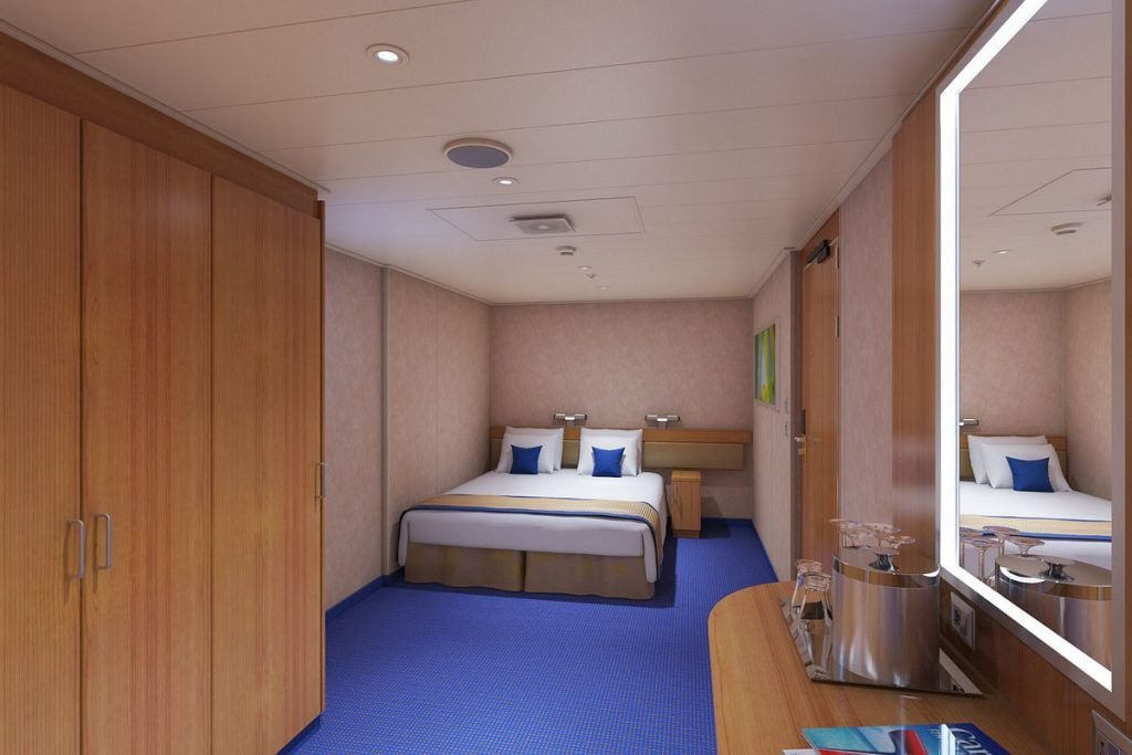 Interior view of a cruise ship cabin with a queen-size bed, blue carpet, wooden accents, and a mirrored wall reflecting the room's layout.