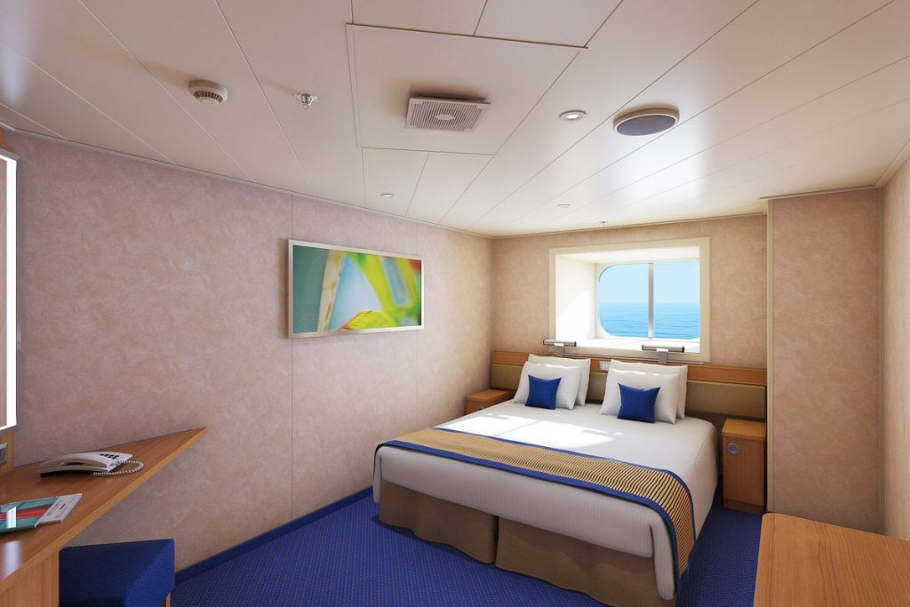 This image shows an OceanView cabin on a Carnival cruise ship. The cabin features a comfortable queen-sized bed adorned with blue and white pillows, positioned beneath a large window offering a view of the ocean. The decor is simple and modern, with light beige walls, blue carpeting, and a wooden desk equipped with a phone and reading materials. A piece of colorful wall art adds a touch of brightness to the room, creating a cozy and inviting atmosphere for guests.